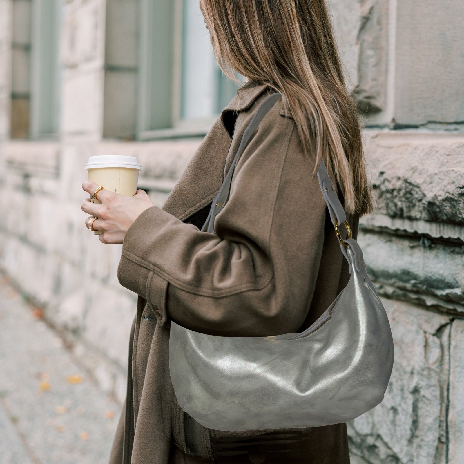 Woman in a brown coat holding a silver bag and a coffee cup, standing against a stone building.