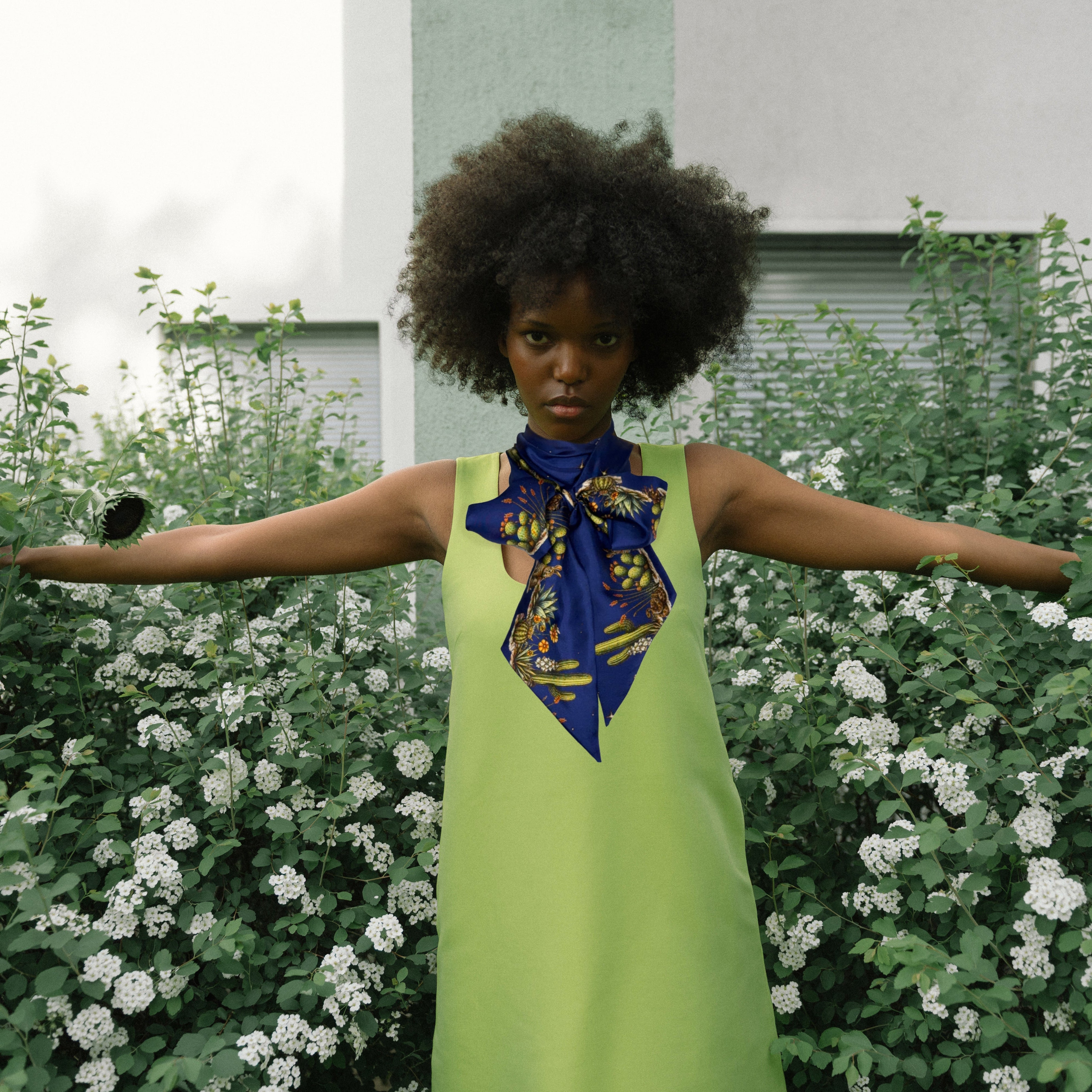 Woman in a green dress with a blue scarf standing in front of greenery