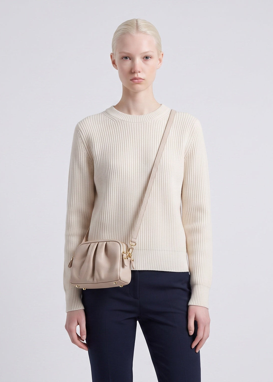 A model stands against a light gray studio background wearing a cream ribbed knit sweater, dark navy pants, and a beige crossbody bag.