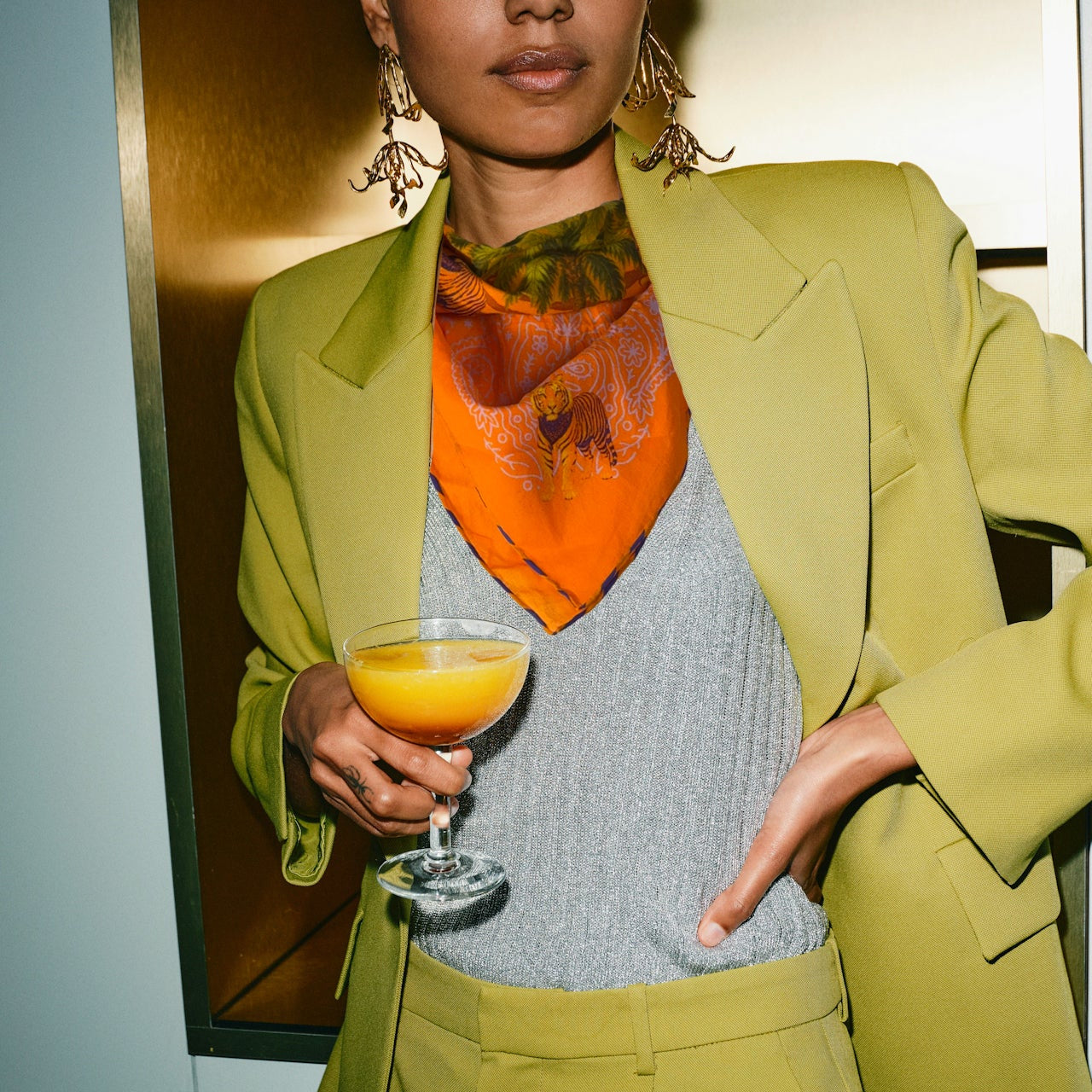 Person in a green suit holding a cocktail with an orange drink and scarf.