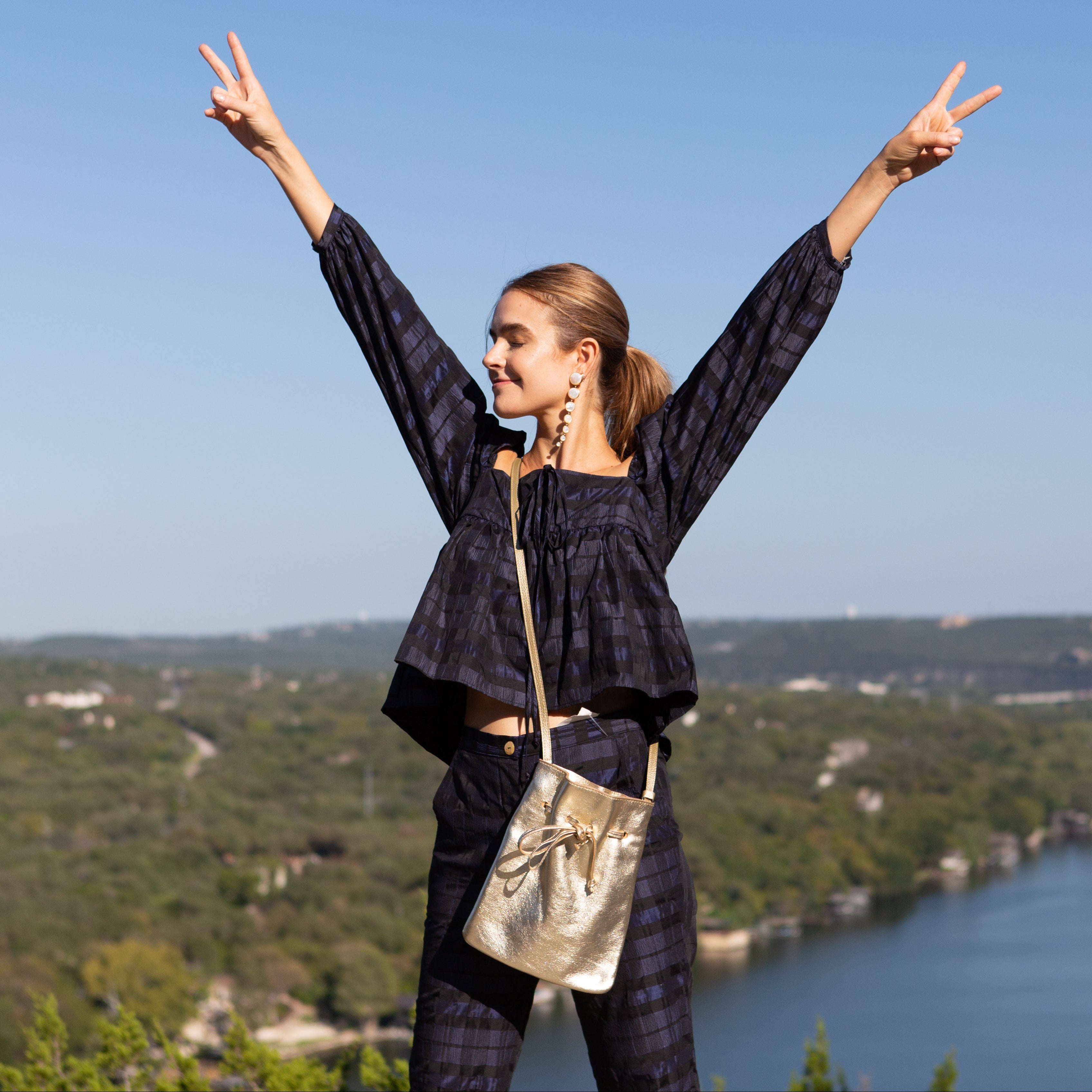 Person standing with arms outstretched over a scenic landscape with a river and hills.