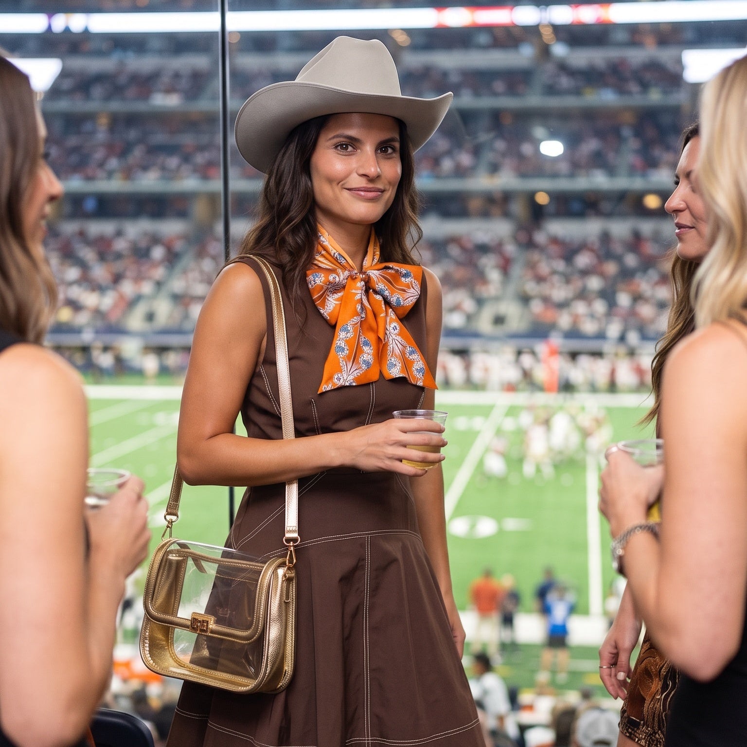 A woman stands in a stadium concession area wearing a brown sleeveless dress, a wide-brim cowboy hat, an orange patterned neck scarf, and brown cowboy boots, with a clear shoulder bag. Other people are partially visible at the sides, and a football field and stadium seating are blurred in the background.
